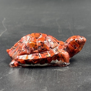 Puede incluir: Una figura de tortuga hecha a mano, que muestra un diseño de mosaico en tonos rojos y marrones. La escultura tiene un acabado liso y brillante, y está sobre un fondo oscuro. La cabeza y las extremidades de la tortuga están claramente definidas.