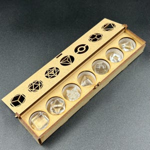 May include: A wooden box with a hinged lid, containing clear, geometric crystal shapes. The lid features cut-out geometric designs. The box is made of light brown wood and sits on a dark surface. The crystals include cubes, pyramids, and other polyhedrons.