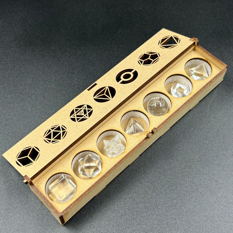 May include: A wooden box with a hinged lid, containing clear, geometric crystal shapes. The lid features cut-out geometric designs. The box is made of light brown wood and sits on a dark surface. The crystals include cubes, pyramids, and other polyhedrons.