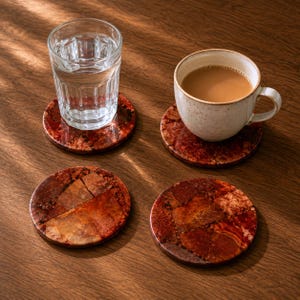 May include: Four circular coasters featuring a mosaic pattern of deep red and brown stone fragments. Two coasters support a clear glass filled with water and a ceramic mug containing a light brown beverage, while two remain empty and visible.