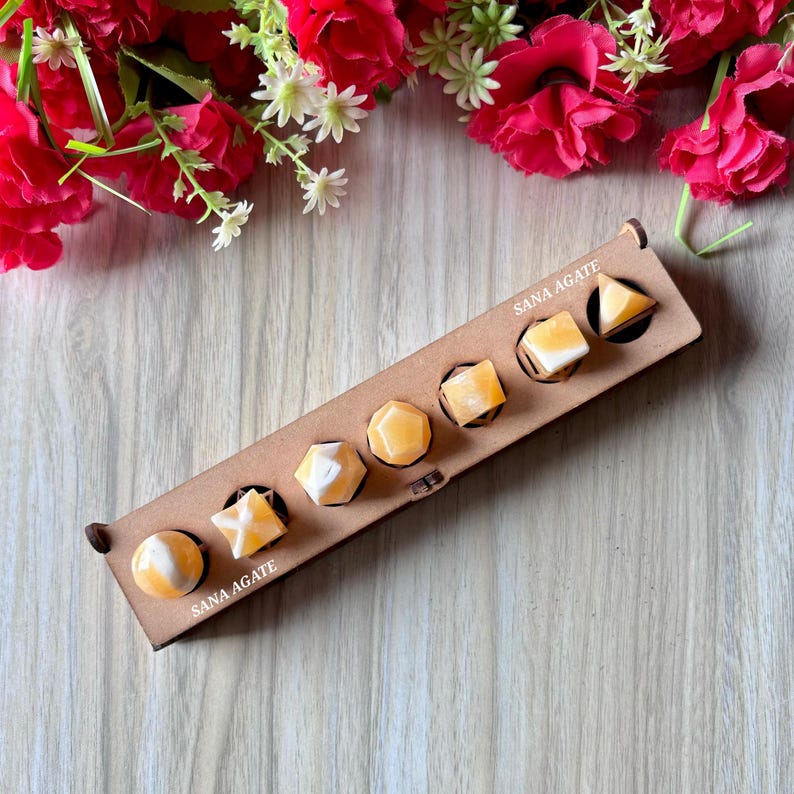 May include: A wooden tray with seven yellow and white gemstone shapes. The tray has the text "SANA AGATE" printed on it. The gemstones are various shapes, including a sphere, star, and triangle. Red flowers and white flowers are in the background.