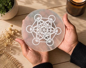 May include: A circular, translucent white selenite charging plate featuring a white engraved Metatron's Cube geometric pattern. The disc has a smooth, polished surface and a flat profile, showcasing the intricate lines of the sacred geometry design.