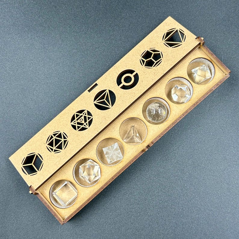 May include: A wooden box with a laser-cut geometric design on the lid, containing clear crystal shapes. The box is open, revealing various crystal forms, including cubes, pyramids, and other polyhedrons. The crystals are set in circular compartments.