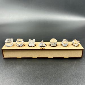 May include: A wooden display box holds seven clear crystal geometric shapes, including cubes, pyramids, and spheres. The crystals are set in individual circular cutouts within the box. The box is made of light brown wood.