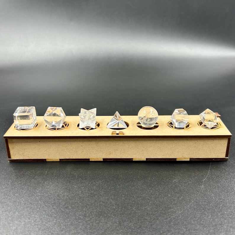 May include: A wooden display box holds seven clear crystal geometric shapes, including cubes, pyramids, and spheres. The crystals are set in individual circular cutouts within the box. The box is made of light brown wood.