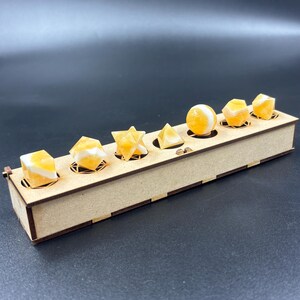 May include: A wooden display box holds seven yellow and white gemstone shapes, including cubes, a star, a pyramid, and a sphere. The gemstones are set in individual circular cutouts within the box, which has a natural wood finish.