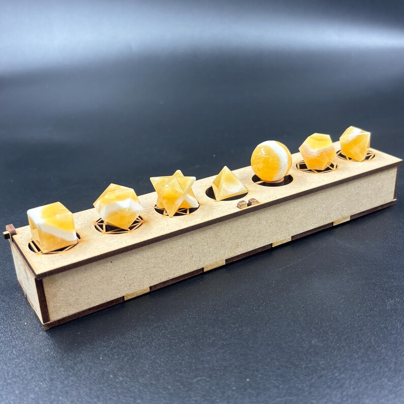 May include: A wooden display box holds seven yellow and white gemstone shapes, including cubes, a star, a pyramid, and a sphere. The gemstones are set in individual circular cutouts within the box, which has a natural wood finish.