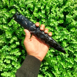Puede incluir: Una punta de flecha de obsidiana negra, también conocida como punta de proyectil, se sostiene en una mano sobre un fondo verde. La punta de flecha mide aproximadamente 25 cm de largo y tiene una punta afilada.