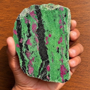 May include: A flat, irregularly shaped slab of zoisite mineral featuring a vibrant green base colour, interspersed with distinct, elongated streaks of deep black hornblende and scattered, irregular patches of bright magenta-coloured ruby crystals.
