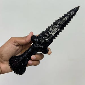 Op de afbeelding: Een zwarte obsidiaan dolk met een gekarteld lemmet en een gedetailleerde handgreep. De dolk wordt in een hand gehouden tegen een witte achtergrond. De tekst "SANA AGATE" is zichtbaar op de handgreep.