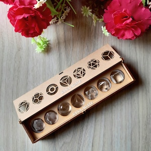 May include: A wooden box with a laser-cut geometric design on the lid, containing clear, faceted crystal stones. The box is labeled "SANA AGATE." Red flowers and greenery are in the background.