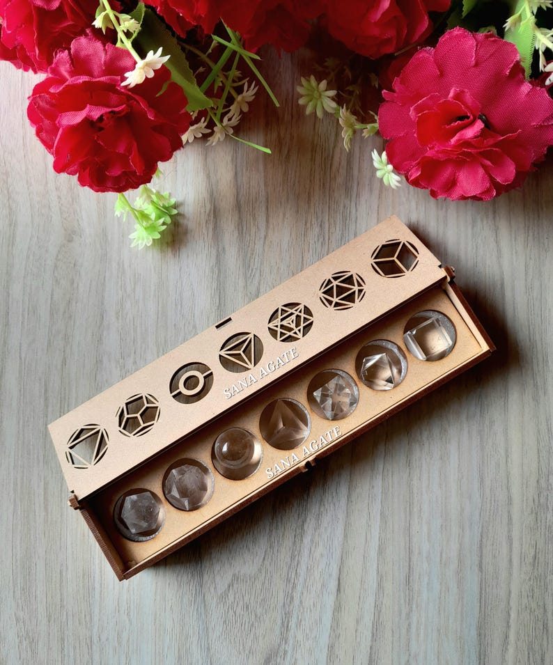 May include: A wooden box with a laser-cut geometric design on the lid, containing clear, faceted crystal stones. The box is labeled "SANA AGATE." Red flowers and greenery are in the background.