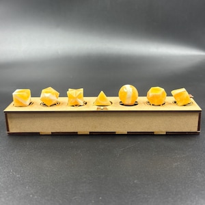 May include: A wooden display box holds seven geometric shapes in various forms. The shapes are a mix of orange and white, with a marbled appearance. The shapes include a cube, a six-sided shape, a folded shape, a pyramid, a sphere, and a rounded shape.
