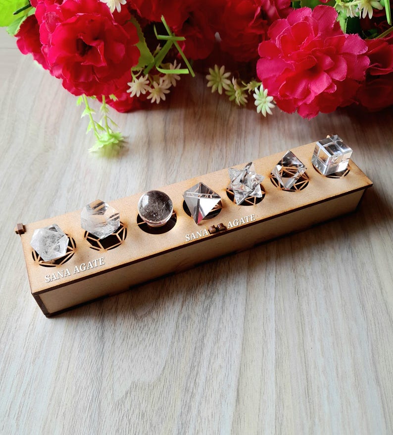May include: A wooden display box with six clear crystals of various shapes, including a sphere, pyramids, and cubes. The crystals are set in decorative holders. The text "SANA AGATE" is printed on the box. Red flowers are in the background.