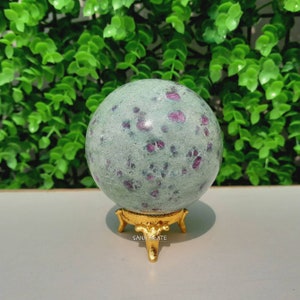May include: A green and pink sphere with red and white flecks, sitting on a gold metal stand with three legs. The stand has the words "Sana Agate" engraved on it.