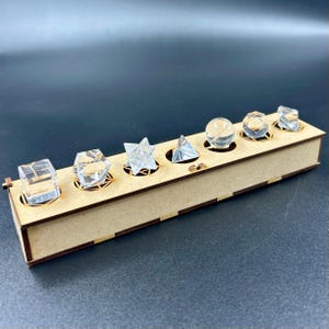 May include: A wooden box with seven clear, geometric shapes displayed in circular cutouts. The shapes include a cube, a hexagon, a star, a pyramid, a sphere, an octahedron, and a rectangular prism. The box is made of light brown wood.