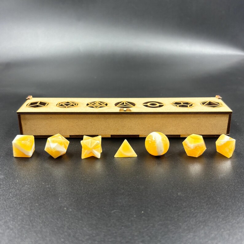 May include: A set of geometric resin dice in various shapes and colors, including cubes, pyramids, and spheres. The dice are yellow and white, and are displayed in a wooden box with decorative cutouts.