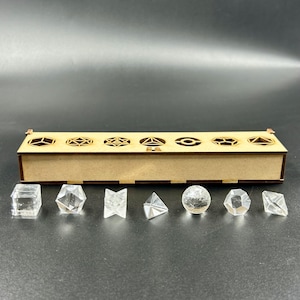 May include: A wooden dice box with geometric cutouts and a set of clear, faceted dice. The dice include a cube, a hexagon, a diamond, a pyramid, a sphere, and other shapes. The box is made of light brown wood.