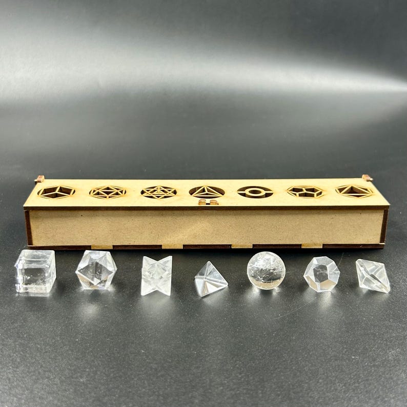 May include: A wooden dice box with geometric cutouts and a set of clear, faceted dice. The dice include a cube, a hexagon, a diamond, a pyramid, a sphere, and other shapes. The box is made of light brown wood.