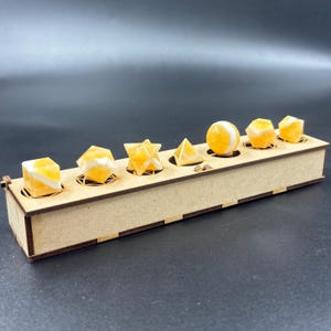 May include: A wooden box with geometric shapes in shades of yellow and white. The shapes include a cube, a diamond, a star, a pyramid, a sphere, and other polyhedrons. The box is made of light-colored wood and has decorative cutouts.