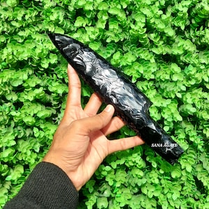 May include: A black obsidian knife with a sharp point and a textured surface. The knife is held in a hand against a green background.