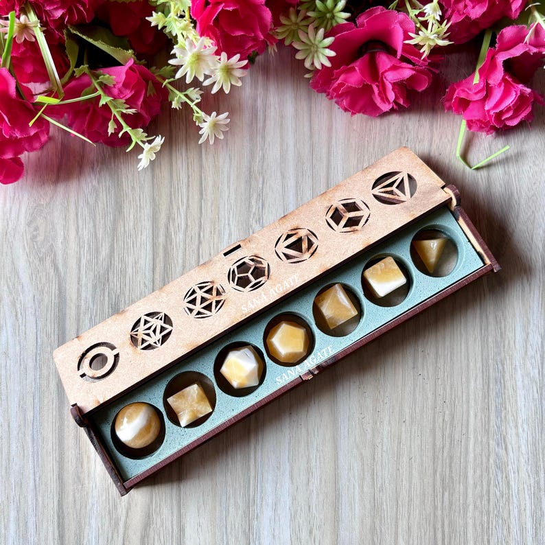 May include: A wooden box with a light blue interior holds seven yellow-orange gemstones of various shapes. The box has a decorative geometric design on the lid and the text "Sana Agate". Pink flowers and greenery are in the background.