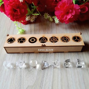 May include: A wooden box with geometric cutouts and the text "SANA AGATE" contains clear crystal shapes. The box is displayed with a backdrop of red flowers and small white flowers. The crystals include a sphere, pyramid, and cube.