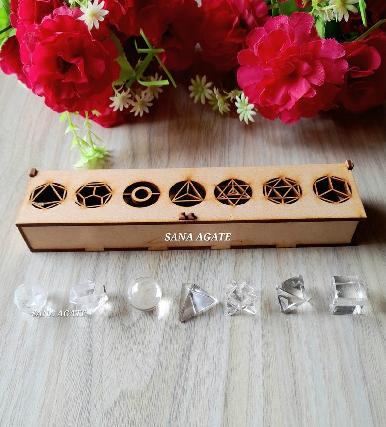 May include: A wooden box with geometric cutouts and the text "SANA AGATE" contains clear crystal shapes. The box is displayed with a backdrop of red flowers and small white flowers. The crystals include a sphere, pyramid, and cube.