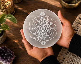 May include: A circular white selenite charging plate featuring a laser-engraved geometric pattern of the flower of life overlaid with seven chakra symbols arranged in a vertical line from the centre to the outer edge of the stone disc.