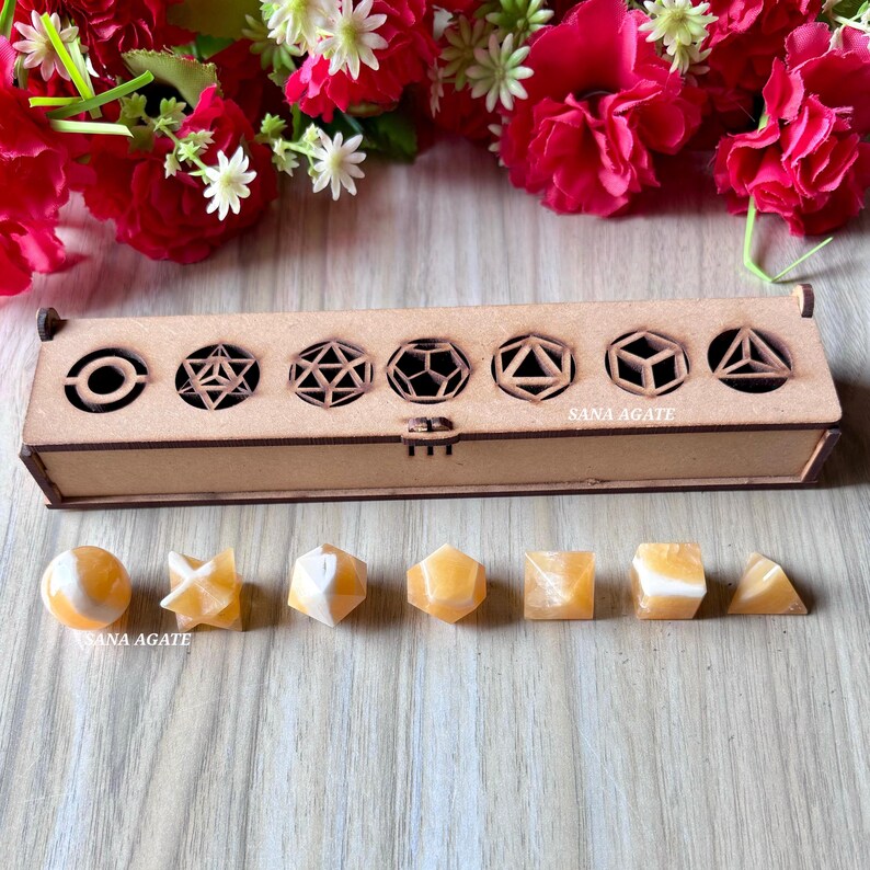 May include: A wooden box with geometric cutouts and a set of orange and white crystal shapes. The box has the text "SANA AGATE" on it. The crystals include a sphere, star, and various polyhedrons.