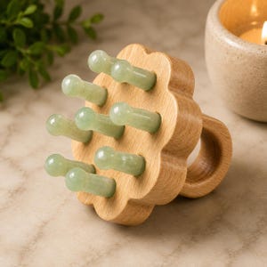 May include: A handheld scalp massager featuring a light-coloured wooden base shaped like a flower with a circular handle on the back. Eight smooth, pale green jade stone prongs are attached to the front of the wooden base for massage application.
