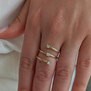 May include: A gold-colored ring with a spiral design, featuring three small, square-cut gemstones. The ring is worn on a hand with manicured nails, set against a white background.