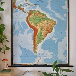 Könnte beinhalten: Eine Vintage-Karte von Südamerika mit der Bezeichnung "SÜDAMERIKA" im Maßstab 1:10.000.000. Die Karte zeigt topografische Details in Grün, Braun und Blau. Die Karte ist mit einem schwarzen Rahmen versehen.