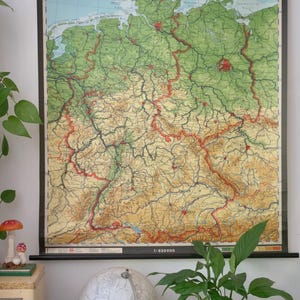 May include: A vintage map of Germany, labeled "DEUTSCHLAND: BUNDESREPUBLIK DEUTSCHLAND UND DDR", with a scale of 1:830000. The map features green, brown, and red hues, mounted on a black frame. A globe and plants are in the foreground.