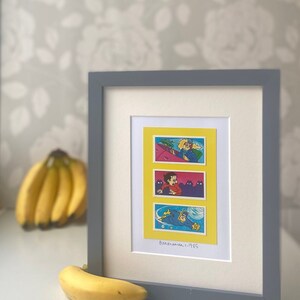 May include: A framed print of three comic book panels featuring a superhero character called "Banana Man" in a yellow, blue, and red costume. The panels are set against a yellow background and the text "Banana Man - 1985" is printed below the panels.