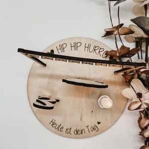 Pu&ograve; includere: Decorazione per compleanno in legno con il testo "Hip Hip Hurray" e "Heute ist dein Tag" (Oggi &egrave; il tuo giorno) in tedesco. La decorazione include una piccola figura di legno e una candela.