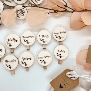 Könnte beinhalten: Holz-Kleiderbügel-Etiketten mit handschriftlichem Text in Deutsch, darunter "Hosen", "Pullis", "Socken", "Strampler", "T-Shirts", "Lange Bodys", "Guize Bodys" und "Schlafes". Die Etiketten sind auf einer weißen Oberfläche mit einer braunen Papiertüte mit dem Text "made with" und einem Herz angeordnet.