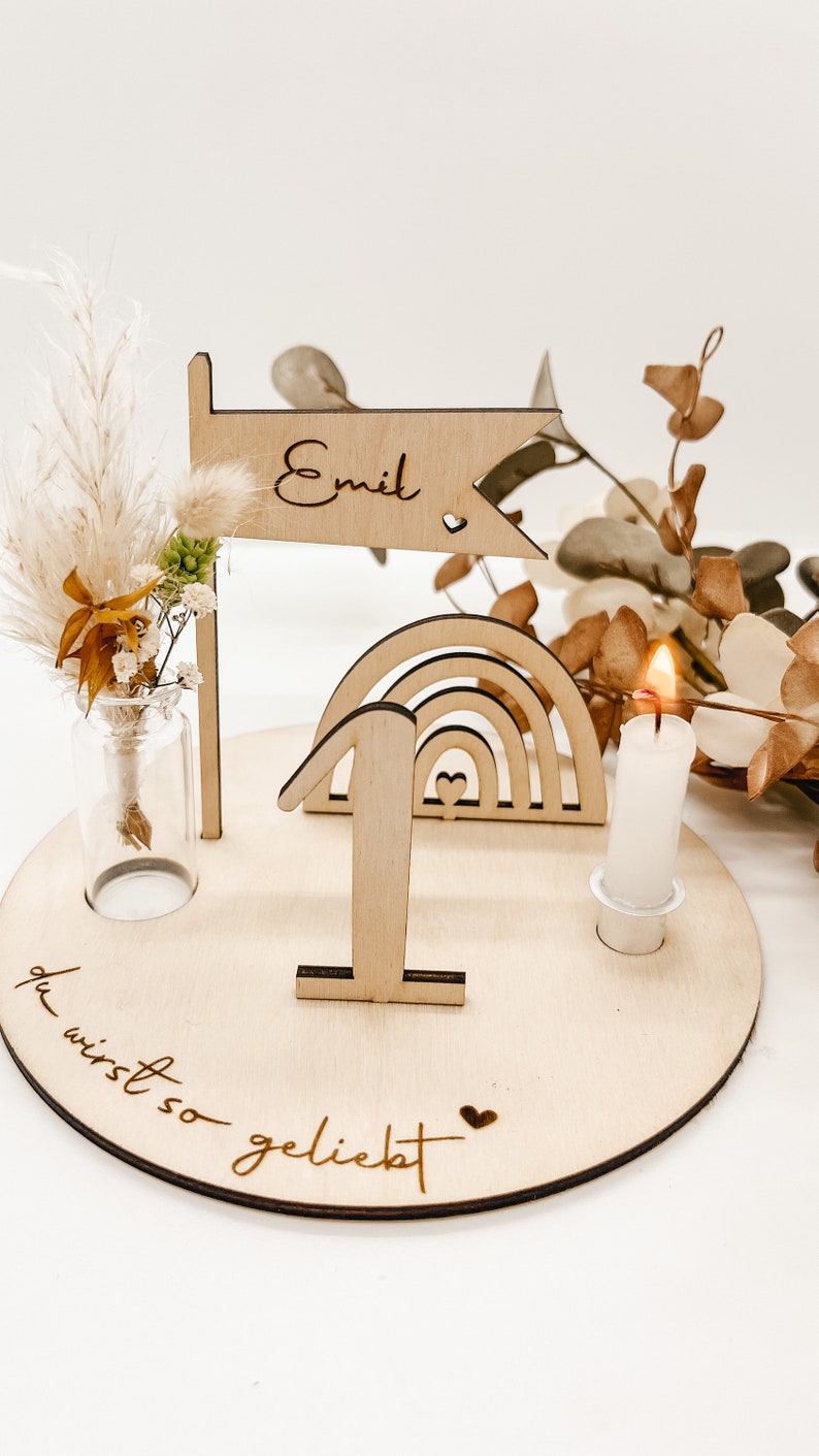 Pu&ograve; includere: Decorazione di compleanno in legno con un arcobaleno, un numero uno, una candela e una bandiera che dice "Emil". La base della decorazione dice "du wirst so geliebt", che si traduce in "sei cos&igrave; amato".