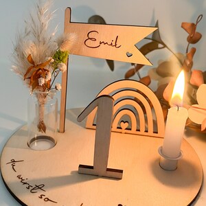 Pu&ograve; includere: Un topper per torta di legno per il primo compleanno con un arcobaleno, un numero uno, un piccolo vaso con fiori secchi, una candela e il testo "du wirst so geliebt" (sei cos&igrave; amato) e "Emil".
