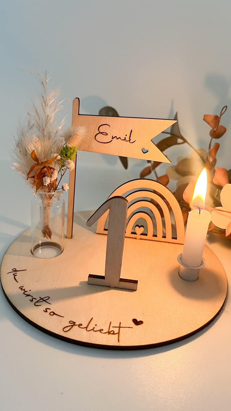 Pu&ograve; includere: Un topper per torta di legno per il primo compleanno con un arcobaleno, un numero uno, un piccolo vaso con fiori secchi, una candela e il testo "du wirst so geliebt" (sei cos&igrave; amato) e "Emil".