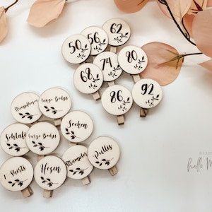 Könnte beinhalten: Holz-Wäscheklammer-Etiketten mit aufgedruckten Zahlen und Wörtern. Die Etiketten dienen zur Organisation von Babykleidung. Die Wörter auf den Etiketten sind "T-Shirts", "Hosen", "Strampler", "Pullis", "Socken", "Schlafi kurze Bodys", "Rumphosen lange Bodys", "50", "56", "62", "68", "74", "80", "86", "92".