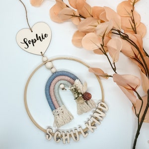 Op de afbeelding: Een houten ring met een regenboog van garen en een houten hart met de naam "Sophie" eraan hangend. De regenboog is versierd met gedroogde bloemen en de ring heeft het woord "Schulkind" erop geschreven in houten letters.