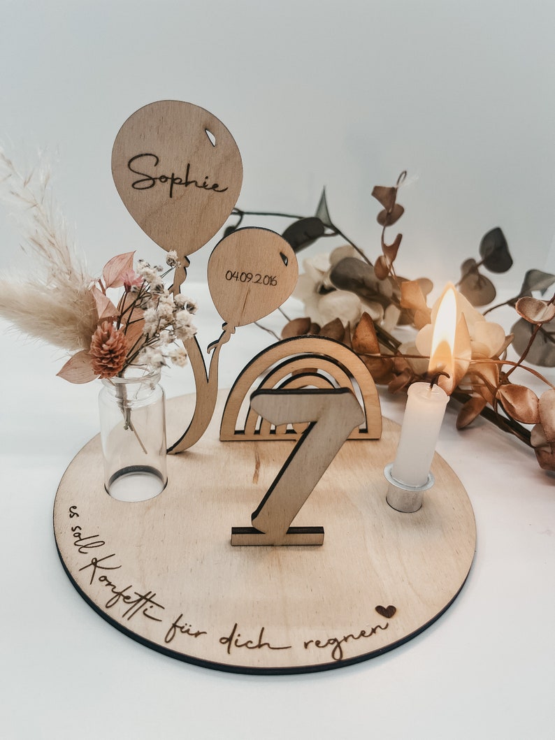 Pu&ograve; includere: Decorazione di compleanno in legno con un arcobaleno, un numero 7, un portacandele e due palloncini. I palloncini sono personalizzati con il nome "Sophie" e la data "04.09.2016". La base della decorazione ha il testo "es soll Konfetti f&uuml;r dich regnen" inciso.