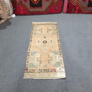 May include: A rectangular area rug featuring a light beige field with a central dark brown floral medallion. The rug includes geometric motifs in muted green and pink, framed by a dark brown border with repeating patterns along the edges.