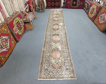Peut inclure: Un long tapis rectangulaire d'aspect délavé et vintage. Le tapis présente un motif central de médaillons roses et bruns répétés, entourés de motifs géométriques dans des tons de beige, de bleu et de vert. Le tapis est posé sur un sol gris.