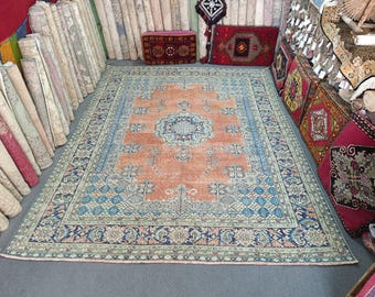 May include: A large, rectangular rug with an intricate design. The rug features a central medallion in shades of blue and peach, surrounded by a border with floral and geometric patterns. The rug is displayed in a shop with other rolled rugs and decorative pillows.