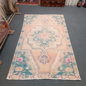 May include: A vintage Turkish rug with a light peach background and a central medallion in shades of teal, pink, and white. The rug has a floral border with a repeating pattern.