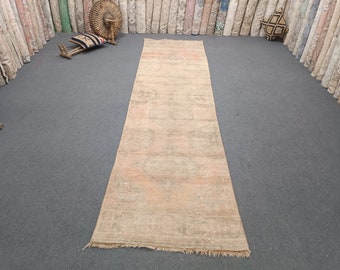 Op de afbeelding: Een lange, rechthoekige vintage-vloerkleed met een vervaagde, versleten uitstraling. Het vloerkleed heeft een centraal ontwerp met geometrische patronen in tinten beige, perzik en crème. Het vloerkleed wordt gepresenteerd op een grijze ondergrond.