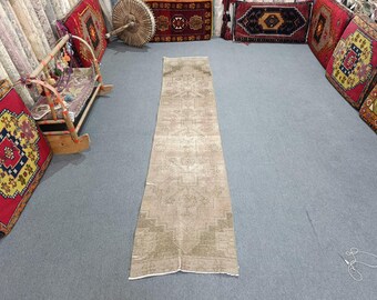 May include: A long, rectangular, beige-colored rug with a faded, distressed appearance. The rug features a central design with geometric patterns and borders. The rug is placed on a gray surface, surrounded by colorful cushions and other rugs.