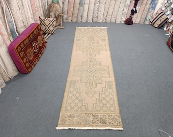 May include: A beige runner rug with a geometric pattern in a darker shade of beige. The rug has a fringe border and is laid out on a gray carpet.